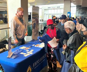 Johns Hopkins Advantage MD stands behind table and provides educational material about our Medicare plans while some visitors read the material.