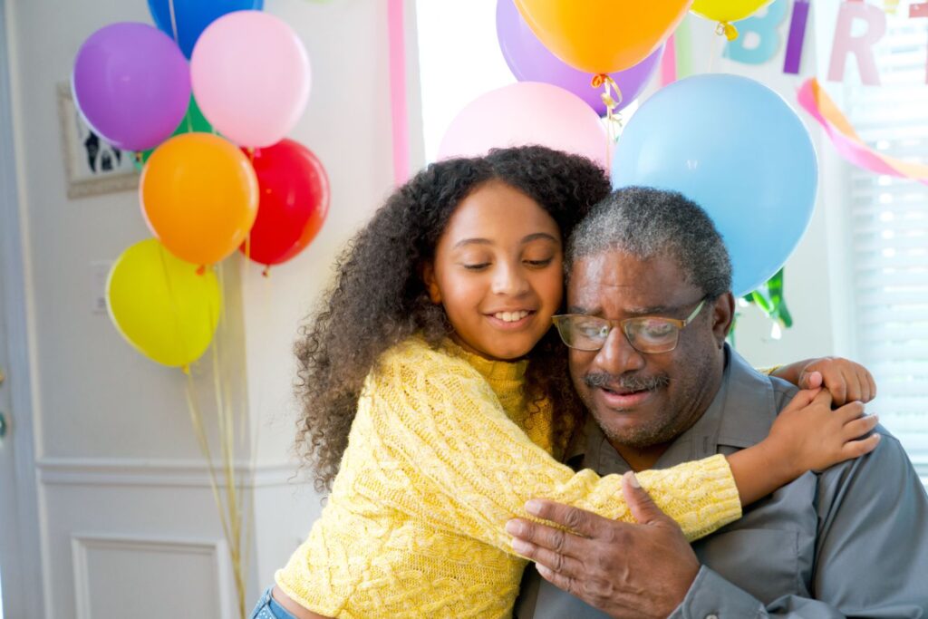 A smiling grandfather wearing glasses hugs his young granddaughter during a colorful birthday celebration with balloons in the background.